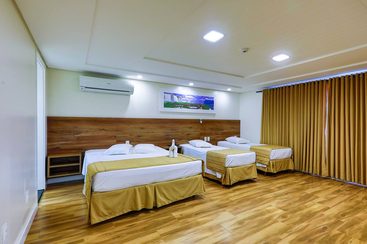 A hotel room with three beds, wooden flooring, an air conditioning unit, a painting on the wall, and a window with curtains.