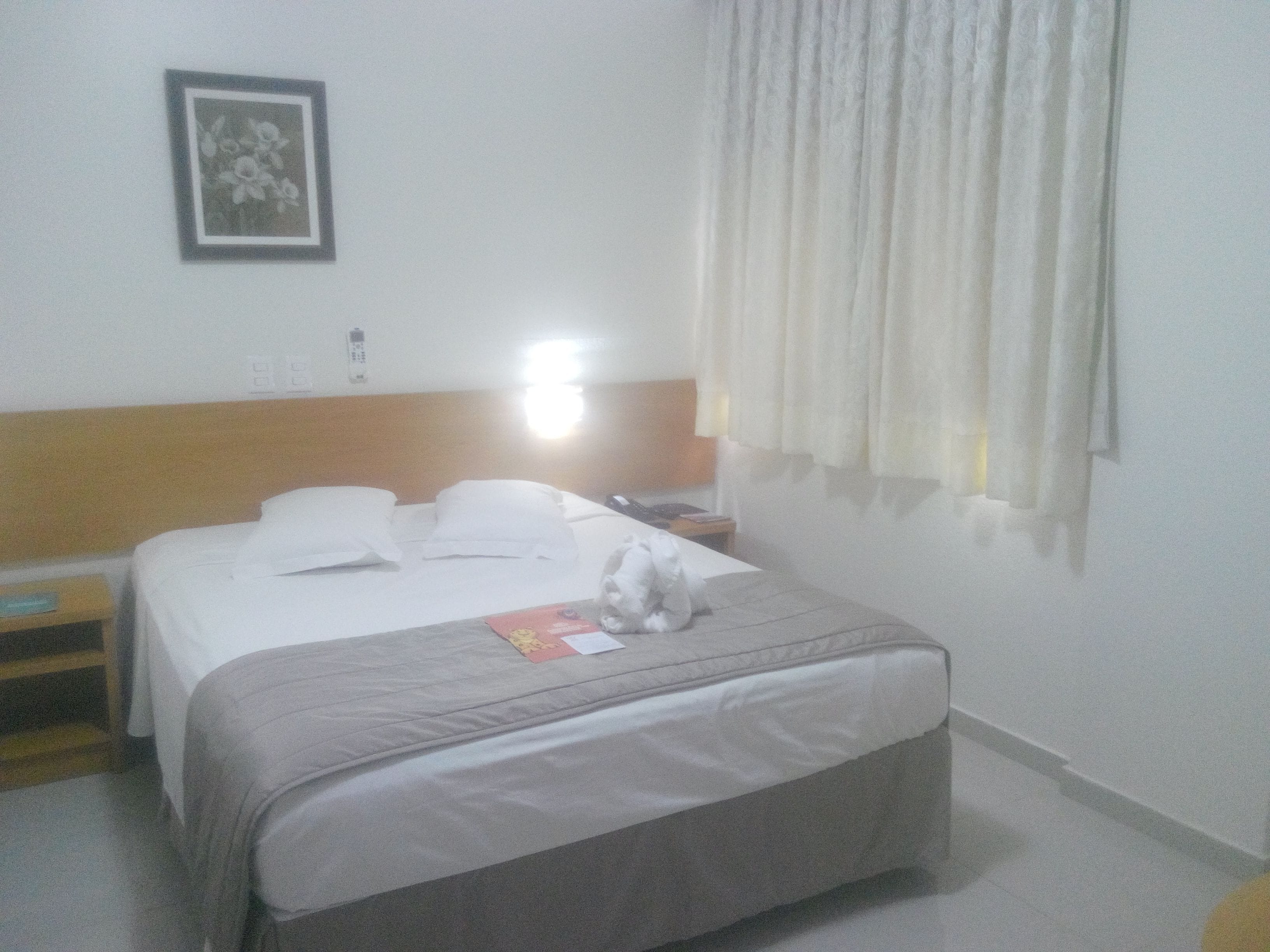 Een hotelkamer met een bed, nachtkastje, een lijst met foto aan de muur en een raam met gordijnen.
