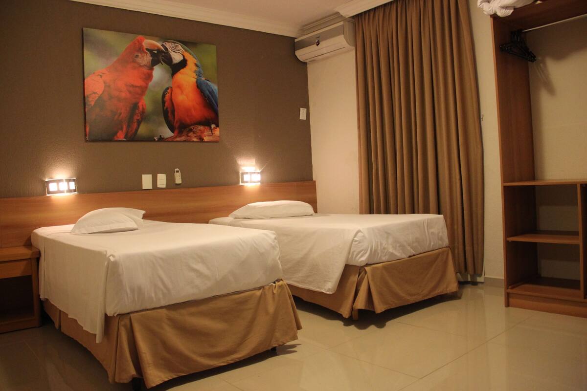 A hotel room with two beds, a painting of parrots, an air conditioning unit, and a wardrobe.