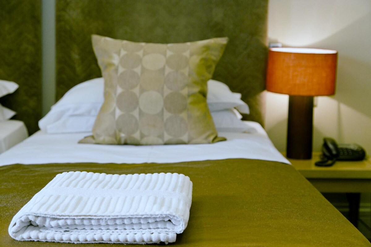 A neatly made bed with a folded white towel, a patterned pillow, and a bedside lamp on a nightstand.