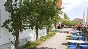 A narrow alleyway with a tree-lined path, a white building with green trim, and outdoor seating with umbrellas. - Hörger Biohotel und Tafernwirtschaft (Kranzberg)