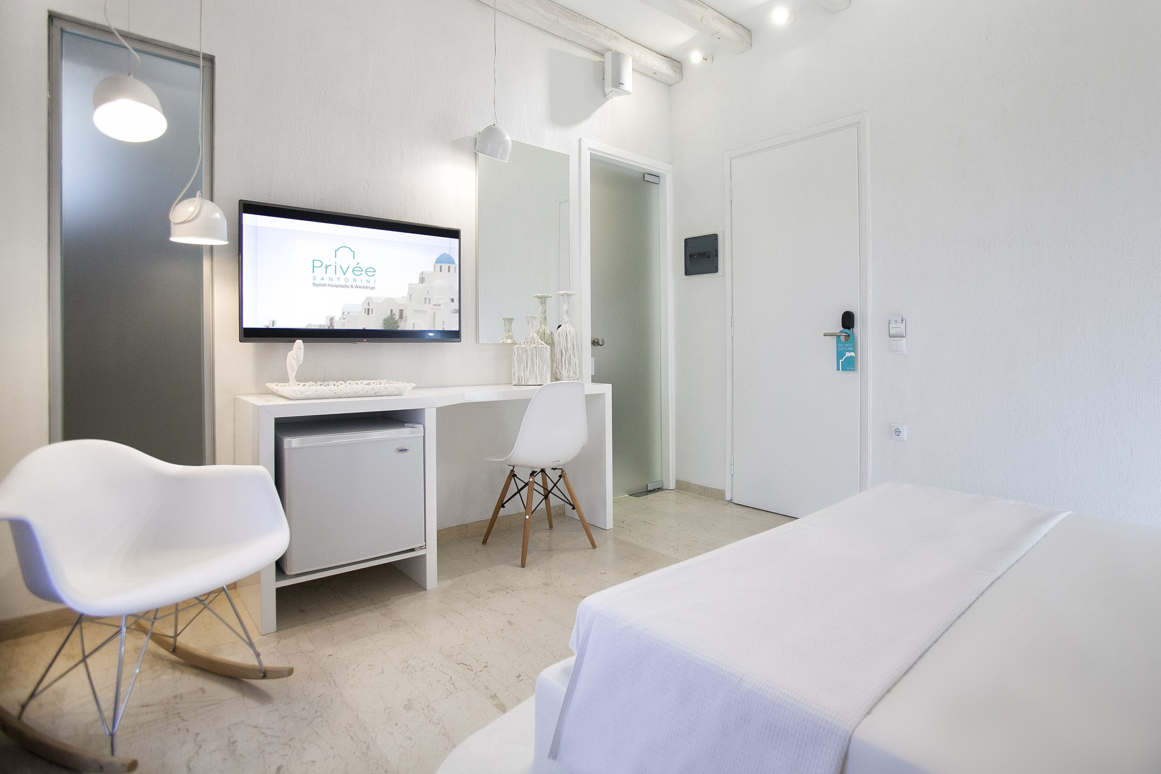 A hotel room with a bed, a desk with a computer, a chair, and a television displaying 'Privée Santorini'.