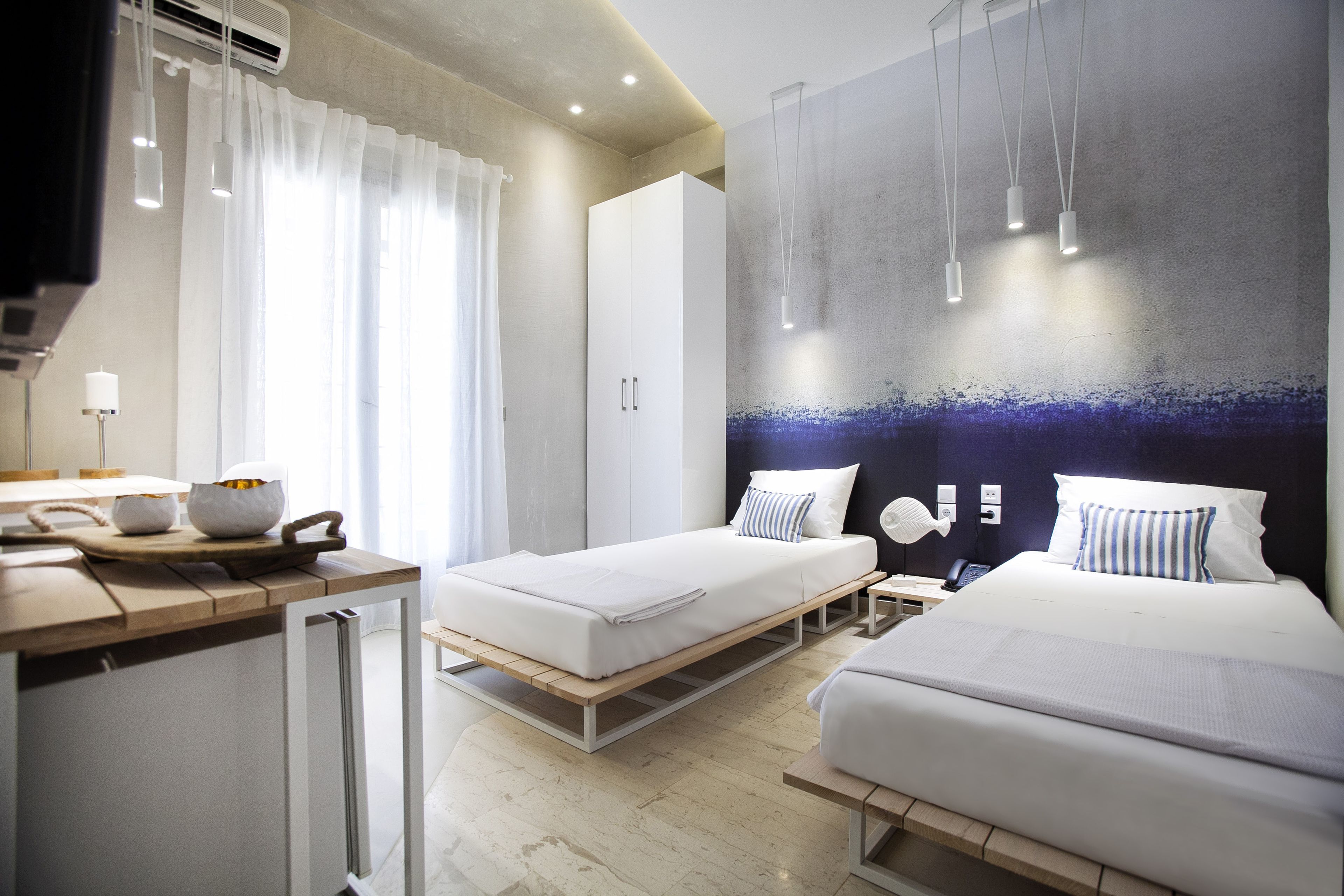 A modern hotel room with two beds, a kitchenette, a large window with curtains, and a wall-mounted light fixture.