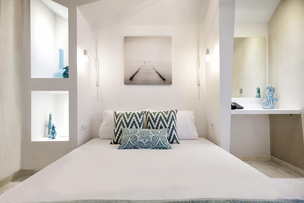 Une chambre moderne avec un grand lit, deux lampes fixées au mur, un tableau au-dessus du lit, et une salle de bain avec un lavabo et un miroir.