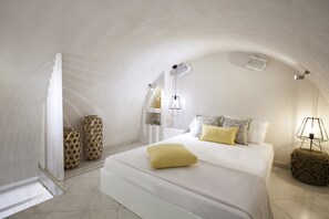 A bedroom with a large bed, a bedside table, a lamp, and a small shelf. - Privee Santorini (Santorini)