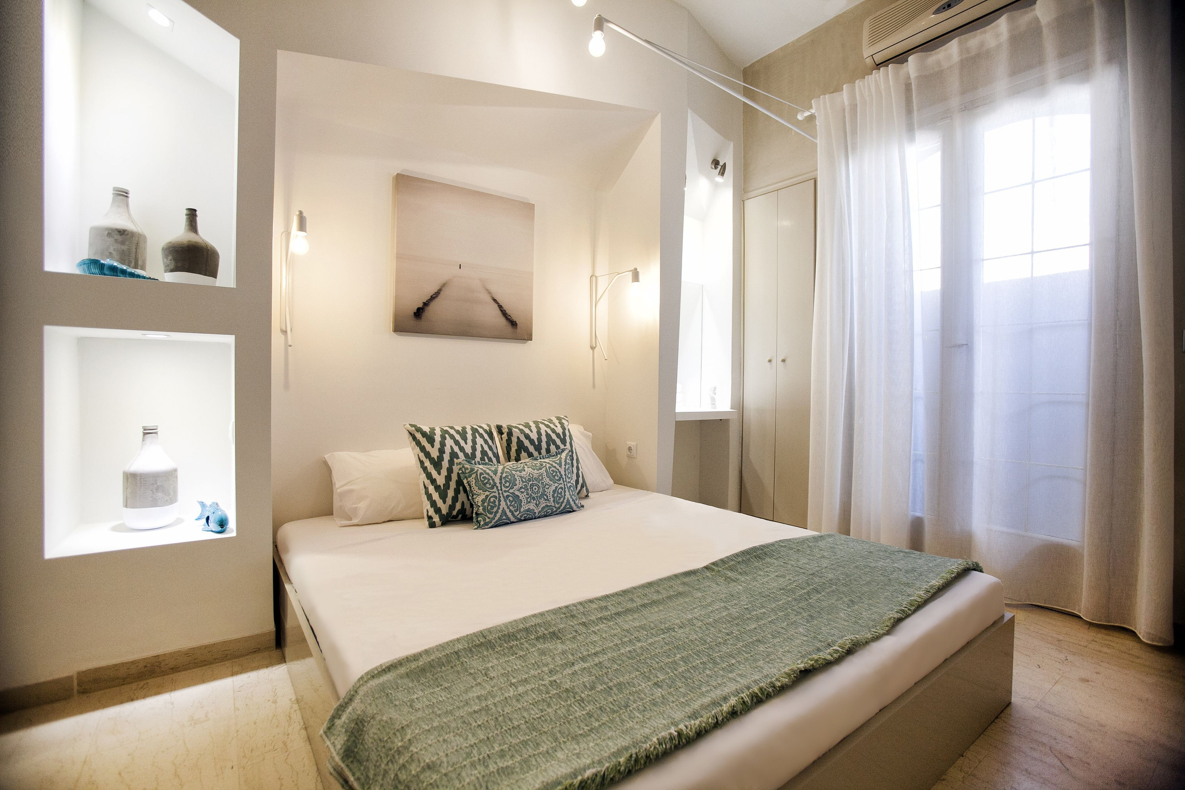 Une chambre moderne avec un grand lit, une table de chevet, une étagère murale agrémentée d’objets décoratifs et une fenêtre avec des rideaux légers.