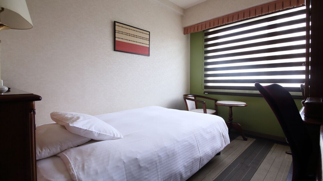 Ein Hotelzimmer mit Bett, kleinem Tisch, Stuhl, Lampe und einem Fenster mit Jalousien.