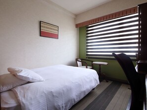 A hotel room with a bed, a chair, a small table, and a window with blinds.