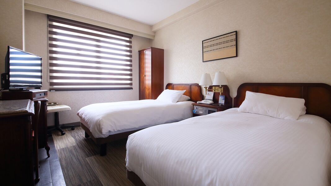 Ein Hotelzimmer mit zwei Betten, einem Schreibtisch mit Fernseher, einem Kleiderschrank und einem Fenster mit Jalousien.