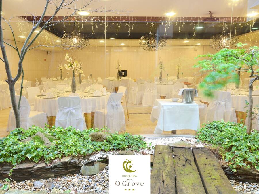 Un salón de eventos de hotel con mesas redondas, manteles blancos y sillas. Hay plantas decorativas y un banco de madera.