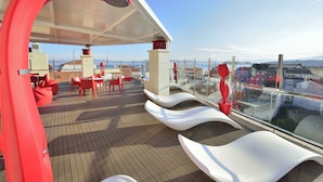 Een modern terras buiten met rode en witte zitplaatsen, een glazen balustrade en uitzicht op de stad.