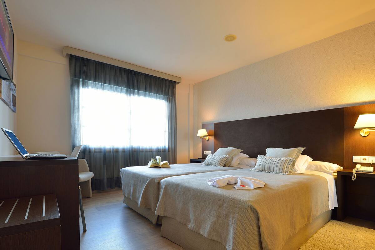 Une chambre d’hôtel avec deux lits, un bureau avec un ordinateur portable, une lampe de chevet et une grande fenêtre avec des rideaux.