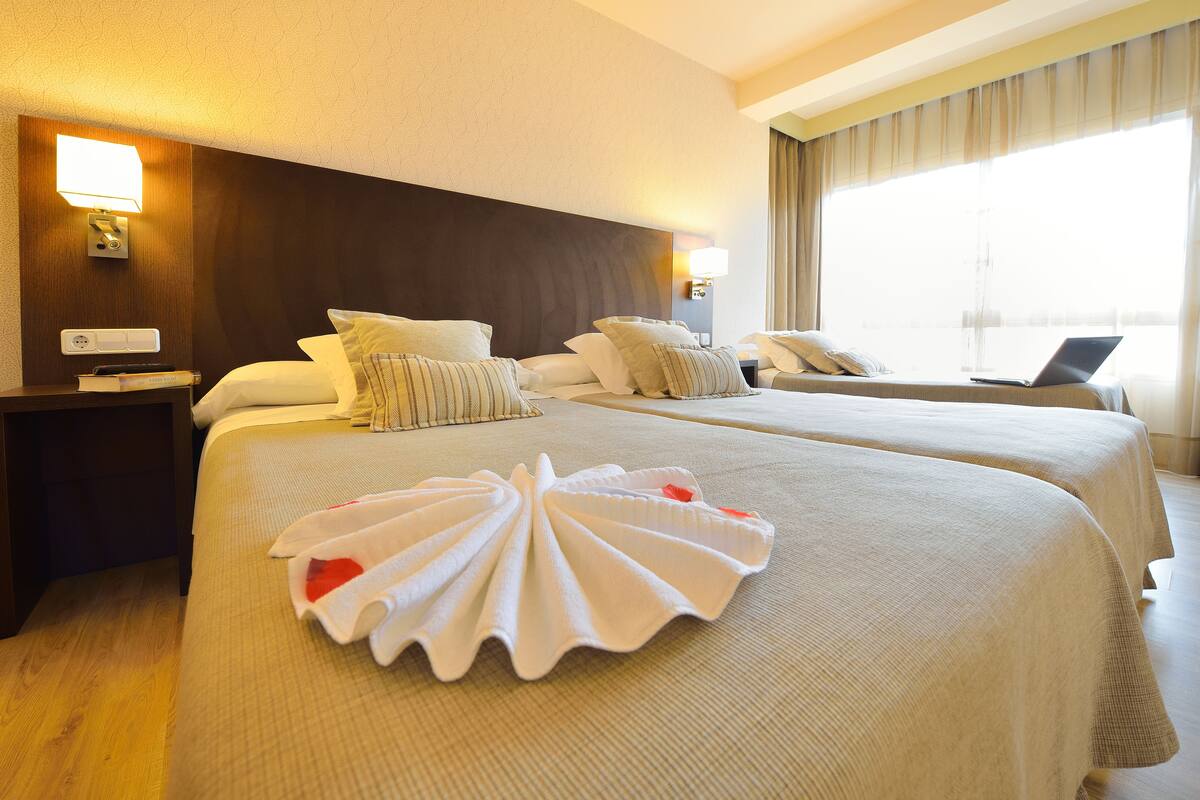 Une chambre d’hôtel avec deux lits, une table de chevet, une lampe, un ordinateur portable posé sur le rebord de la fenêtre et une serviette décorative sur le lit.