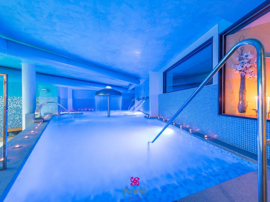 Une piscine intérieure avec un plafond éclairé en bleu, un mur en verre orné d’un vase décoratif et un sol carrelé.