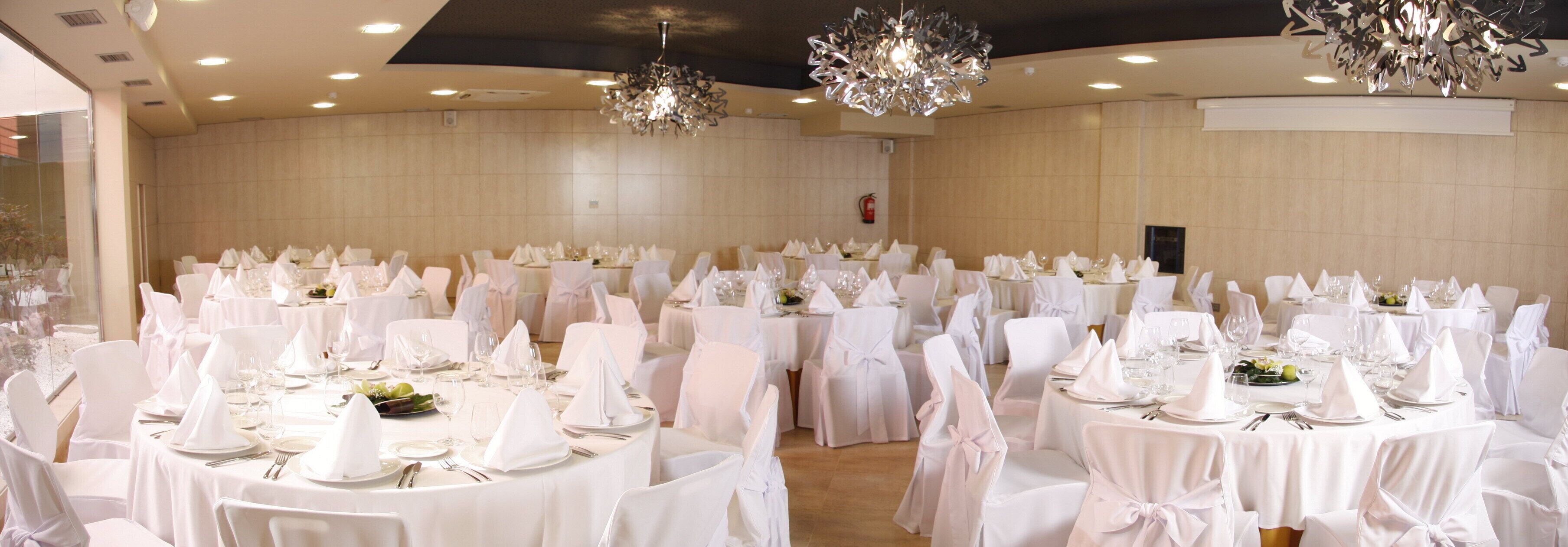 A banquet hall with round tables set for a formal event, featuring white tablecloths and chairs, and decorative crystal chandeliers.