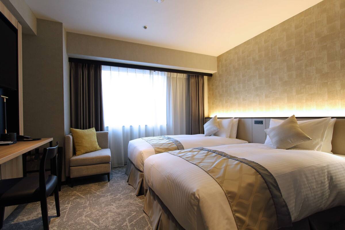 Habitación de hotel con dos camas, un escritorio, una silla y un amplio ventanal con cortinas.