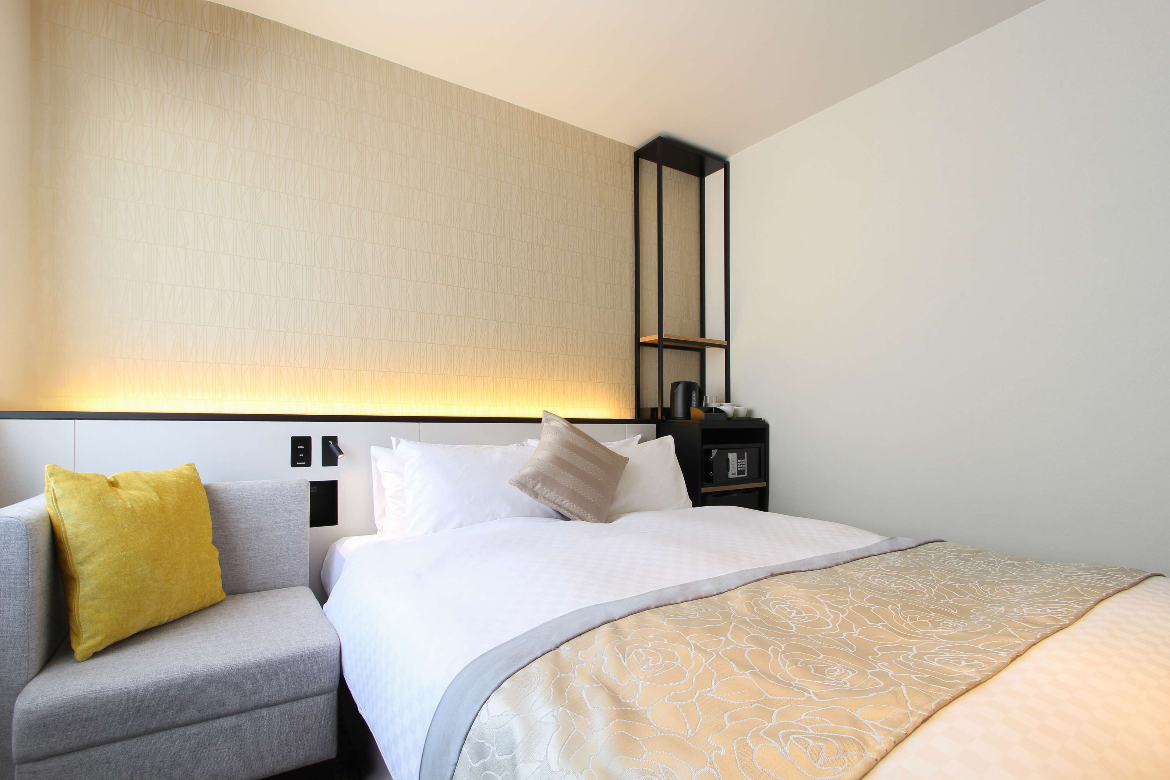 A hotel room with a bed, a grey armchair, a yellow pillow, a bedside table with a lamp, and a wall-mounted shelf.