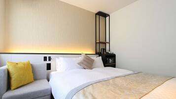 A hotel room with a bed, a grey armchair, a yellow pillow, a bedside table with a lamp, and a wall-mounted shelf.