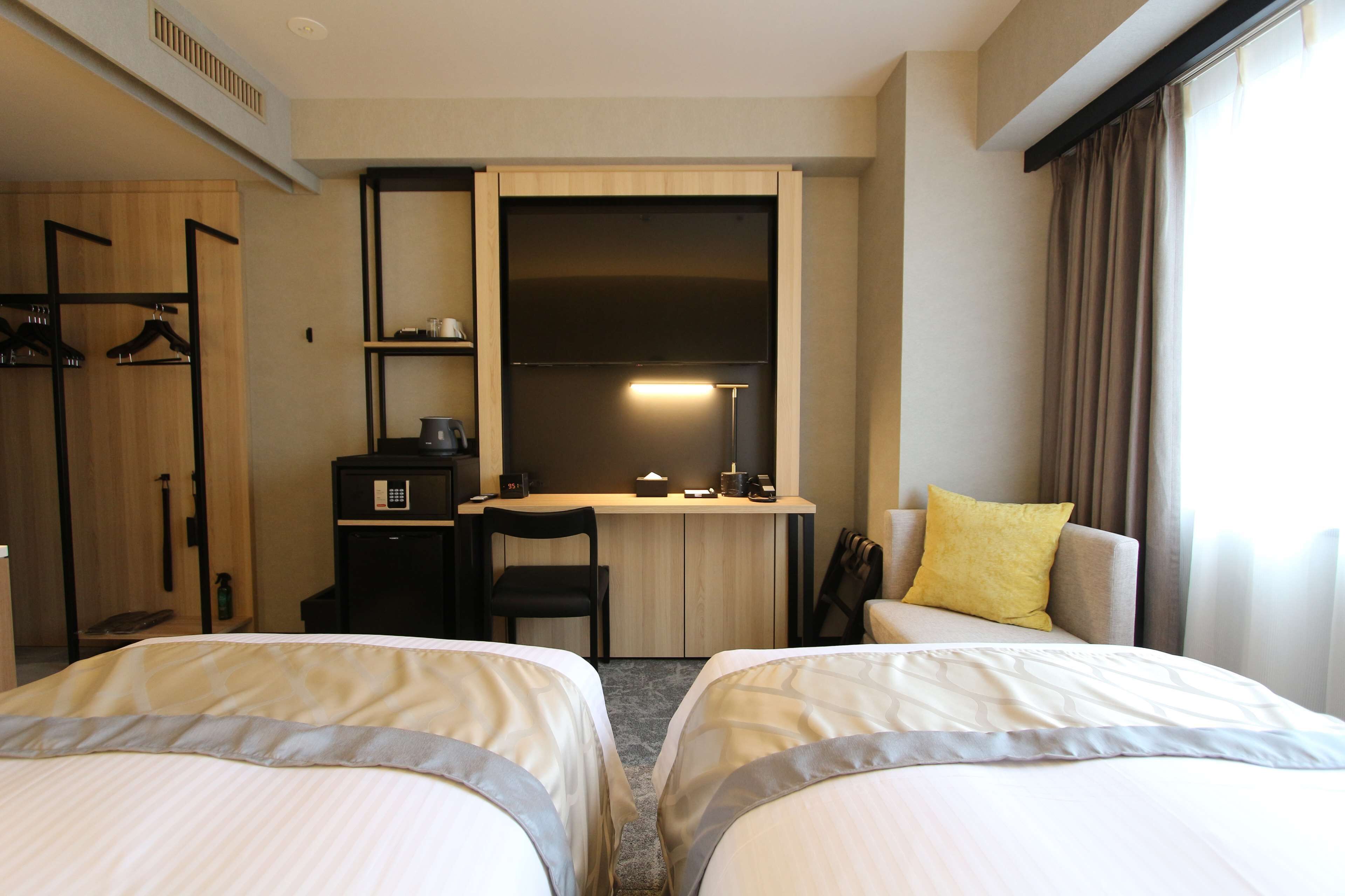 A hotel room with two beds, a desk, a chair, a wardrobe, and a window with curtains.