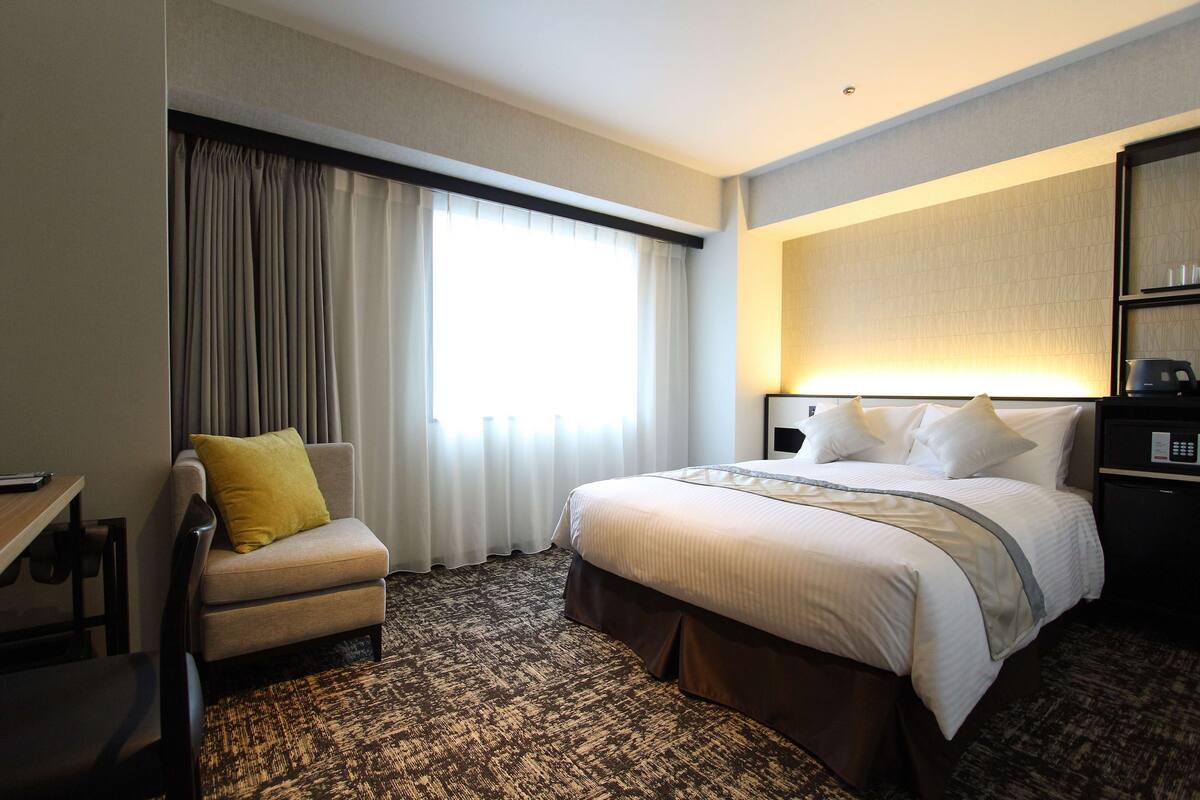 Habitación de hotel con cama, una silla, escritorio y un ventanal con cortinas.