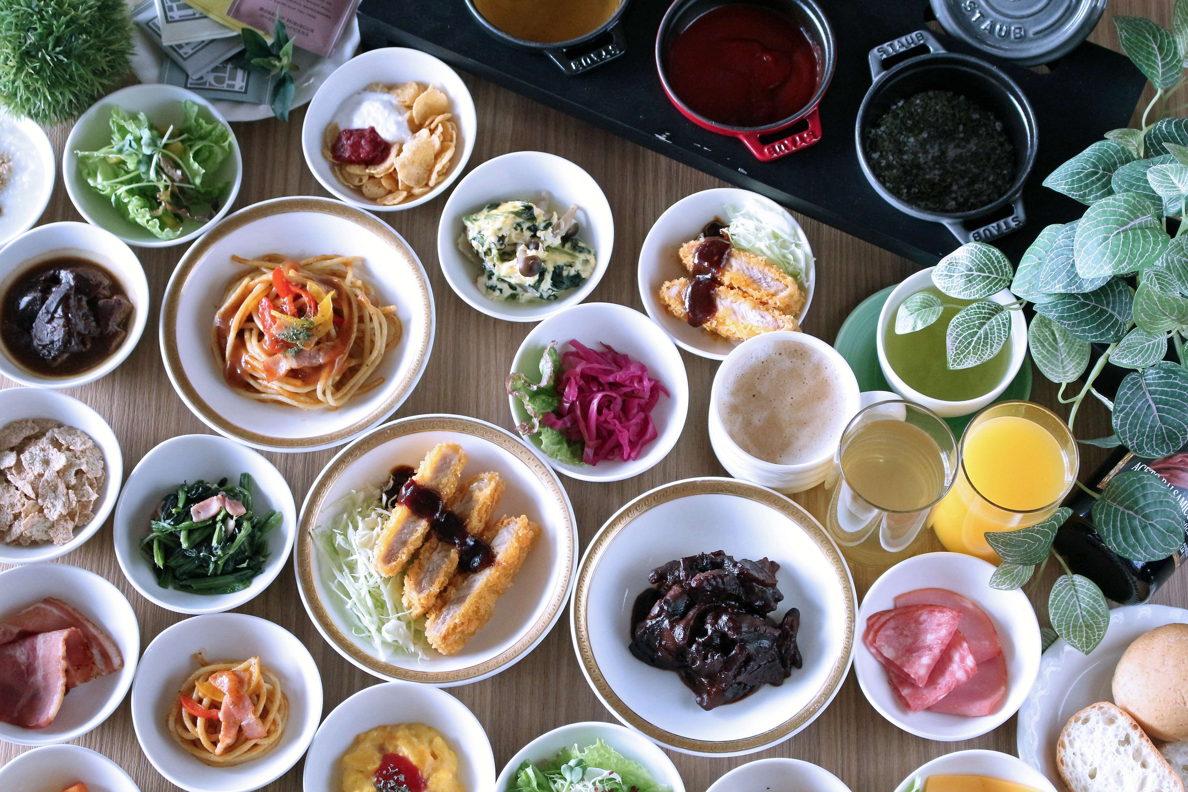 A variety of dishes and drinks on a wooden table, including salads, pasta, and meat dishes, along with two glasses of orange juice and a cup of green tea.