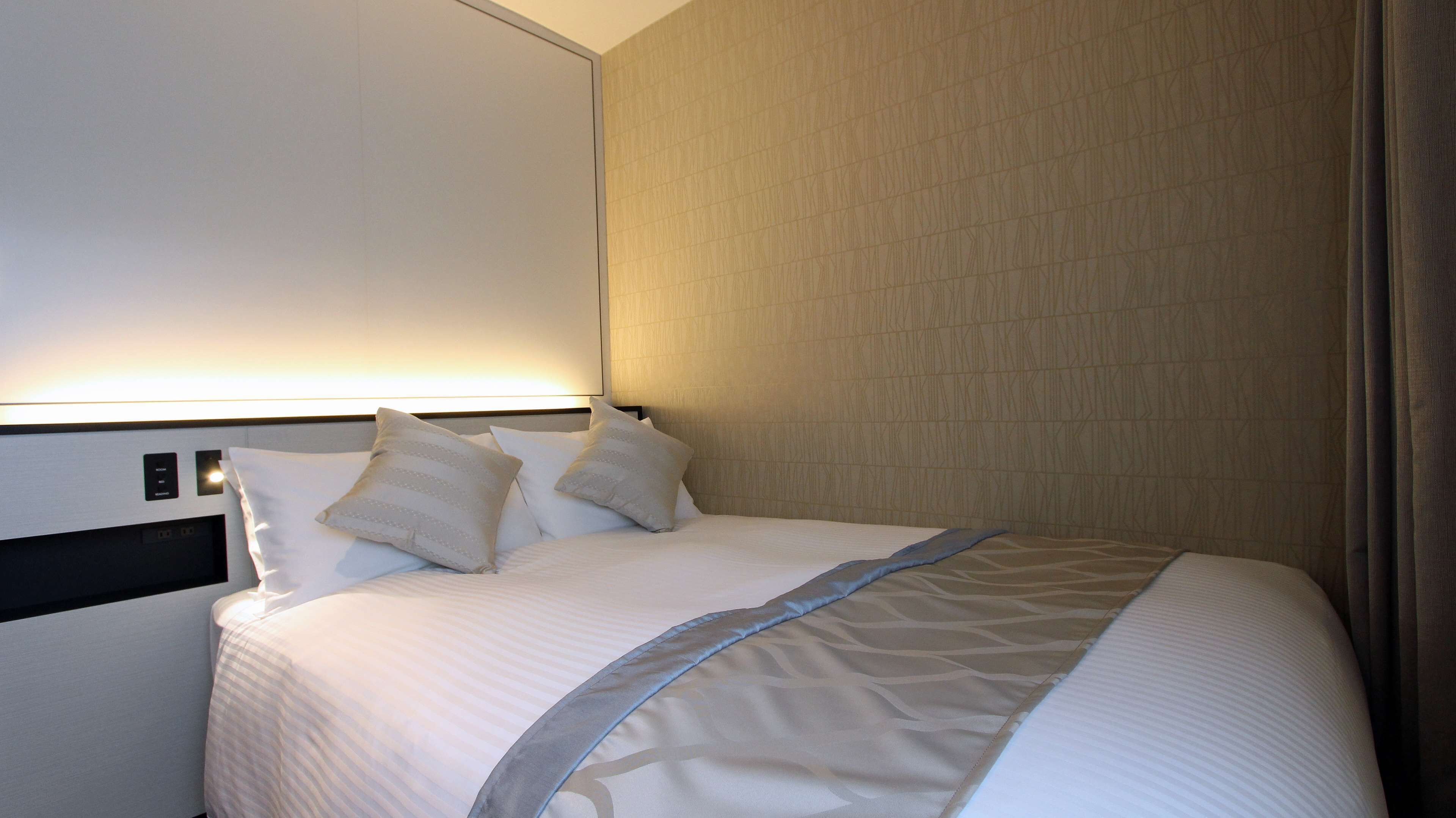 Ein Hotelzimmer mit einem ordentlich bezogenem Bett, einem Kopfteil mit Bedienelementen und einer Wand mit einem geometrischen Muster.