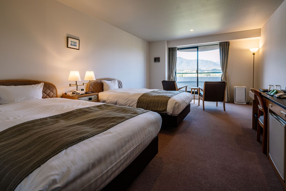 A hotel room with two beds, a desk, a chair, and a view of the outdoors.