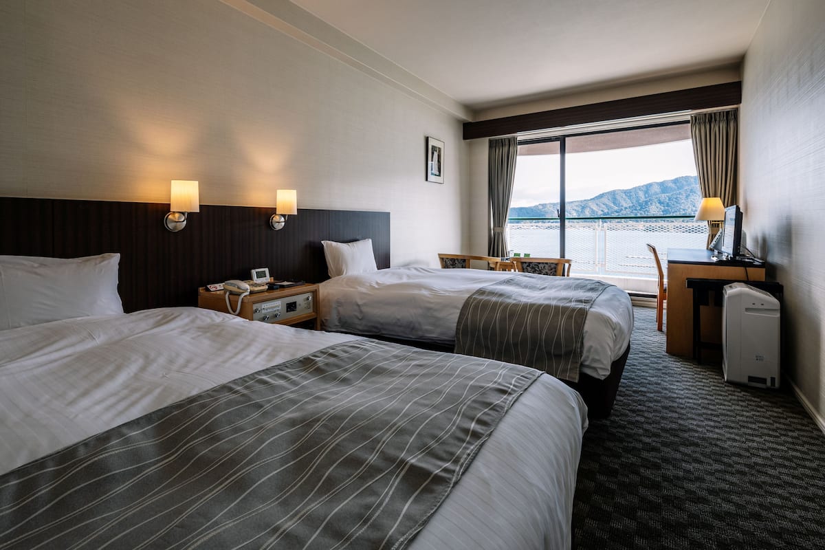 A hotel room with two beds, a desk, a chair, a lamp, and a view of the water.
