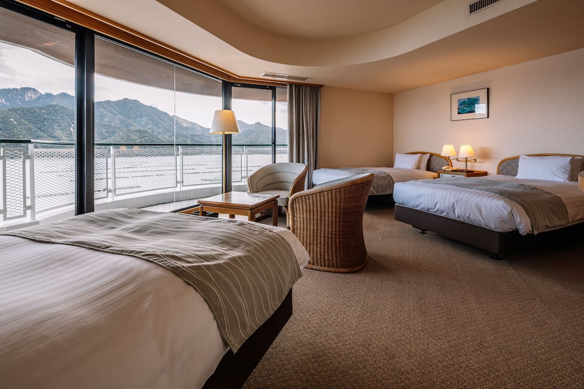 A hotel room with two beds, a chair, a table, and a large window with a view of mountains.