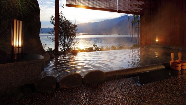 A hot spring bath area with a view of a lake and mountains, wooden architecture, and a stone pathway. - Aki Grand Hotel & Spa (Hatsukaichi)
