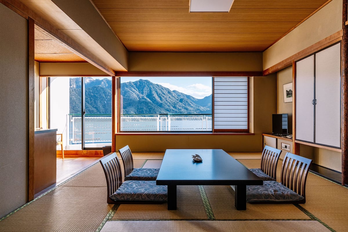 A traditional Japanese room with tatami flooring, a low table, and chairs, a sliding door, and a view of mountains.
