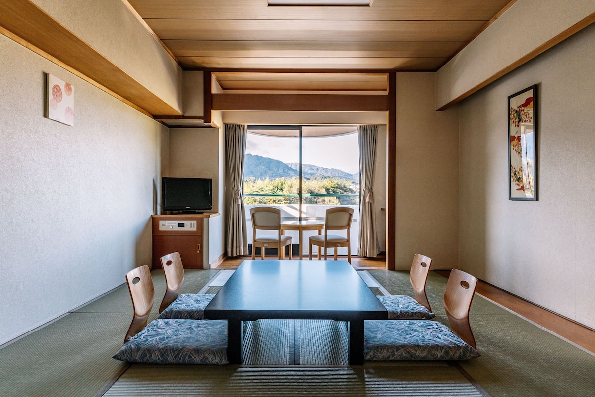 A room with a long table, chairs, a TV, and a view of mountains through large windows.