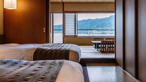 A hotel room with a bed, a bedside table, a window with a view of mountains, and a balcony with a table and chairs. - Aki Grand Hotel & Spa (Hatsukaichi)