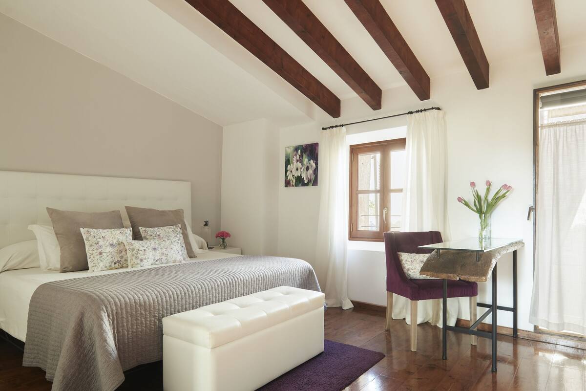 Een slaapkamer met een bed, nachtkastje, stoel, tafel en raam met gordijnen.