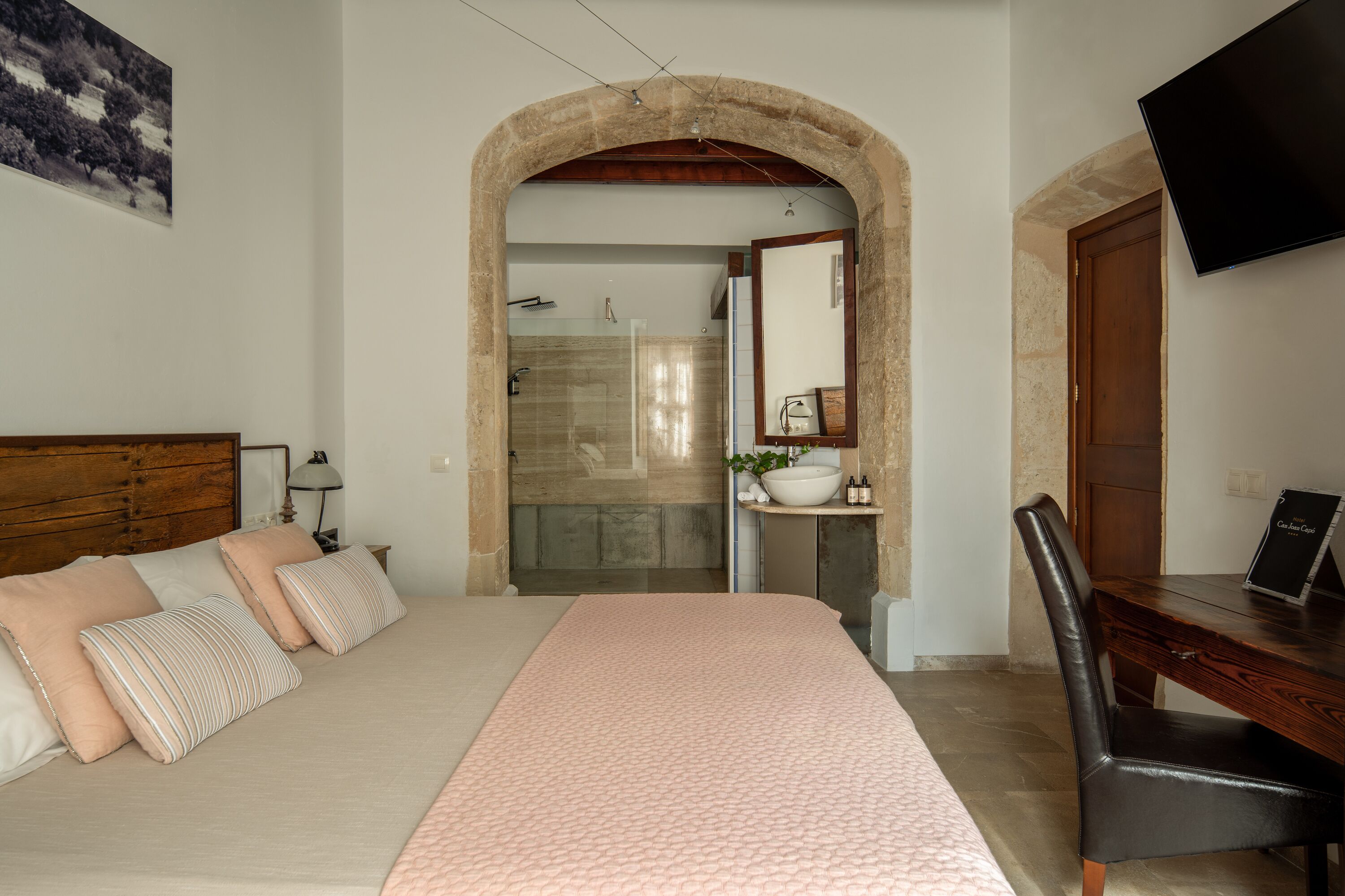 Une chambre à coucher comprenant un lit, un bureau, une chaise, un téléviseur, et une salle de bain visible à travers une porte en arc de cercle.