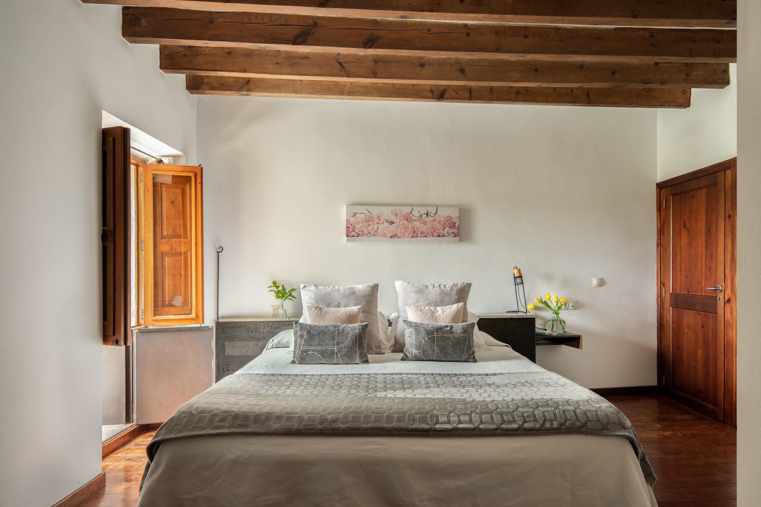 Un dormitorio con techo de madera, una cama con ropa de cama gris, una mesita de noche con un jarrón de flores y una puerta de madera.