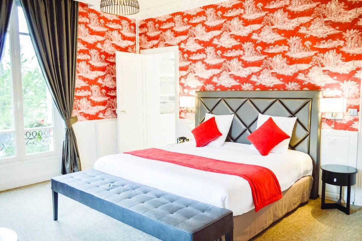 A hotel room with a large bed, a grey upholstered bench, a white wardrobe, and a red patterned wallpaper.
