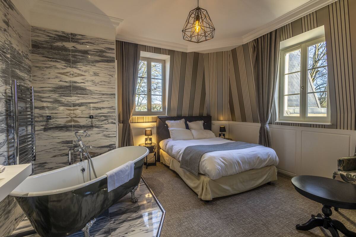 A modern hotel room with a marble bathroom, a large bed, and a view of trees through the windows.