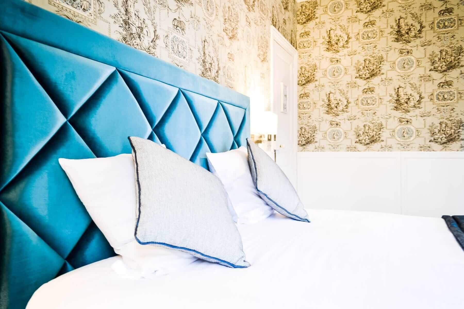 A bed with a blue headboard and two pillows, a patterned wallpaper, and a white door.