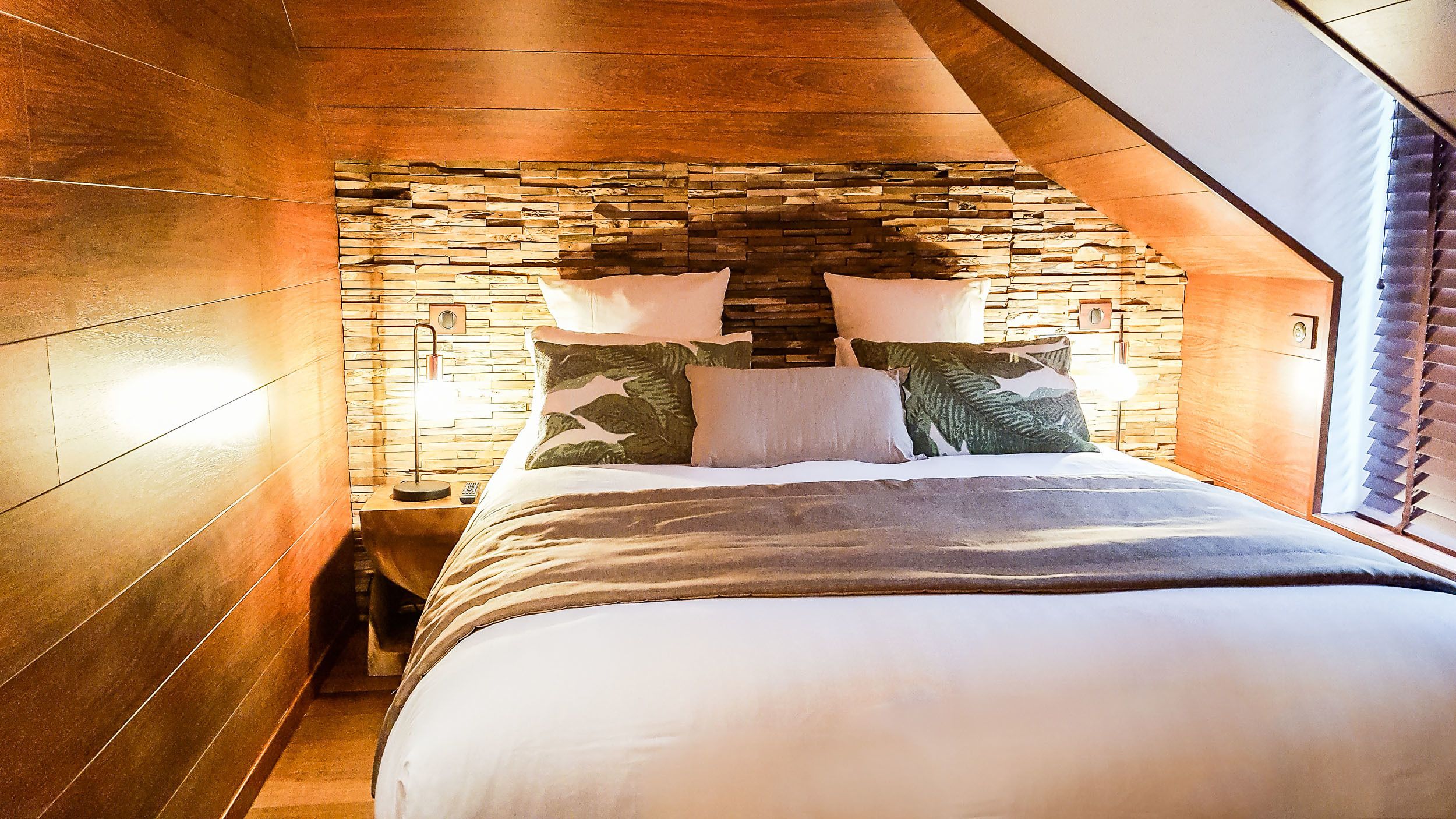 Un dormitorio con pared de piedra, cama con ropa de cama blanca y dos lámparas de noche.