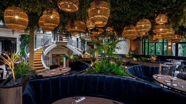Un restaurante con mesas redondas, asientos tapizados en azul oscuro y lámparas de mimbre colgantes.