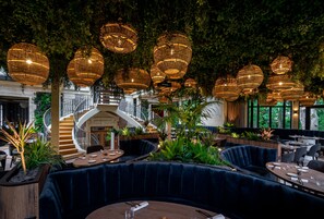 Un restaurant avec des tables rondes, des sièges rembourrés en tissu bleu foncé et des lampes suspendues en osier.
