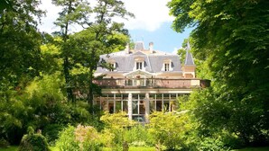 Una casa grande e histórica con una imponente torre central, rodeada de abundante vegetación y un jardín cuidado.