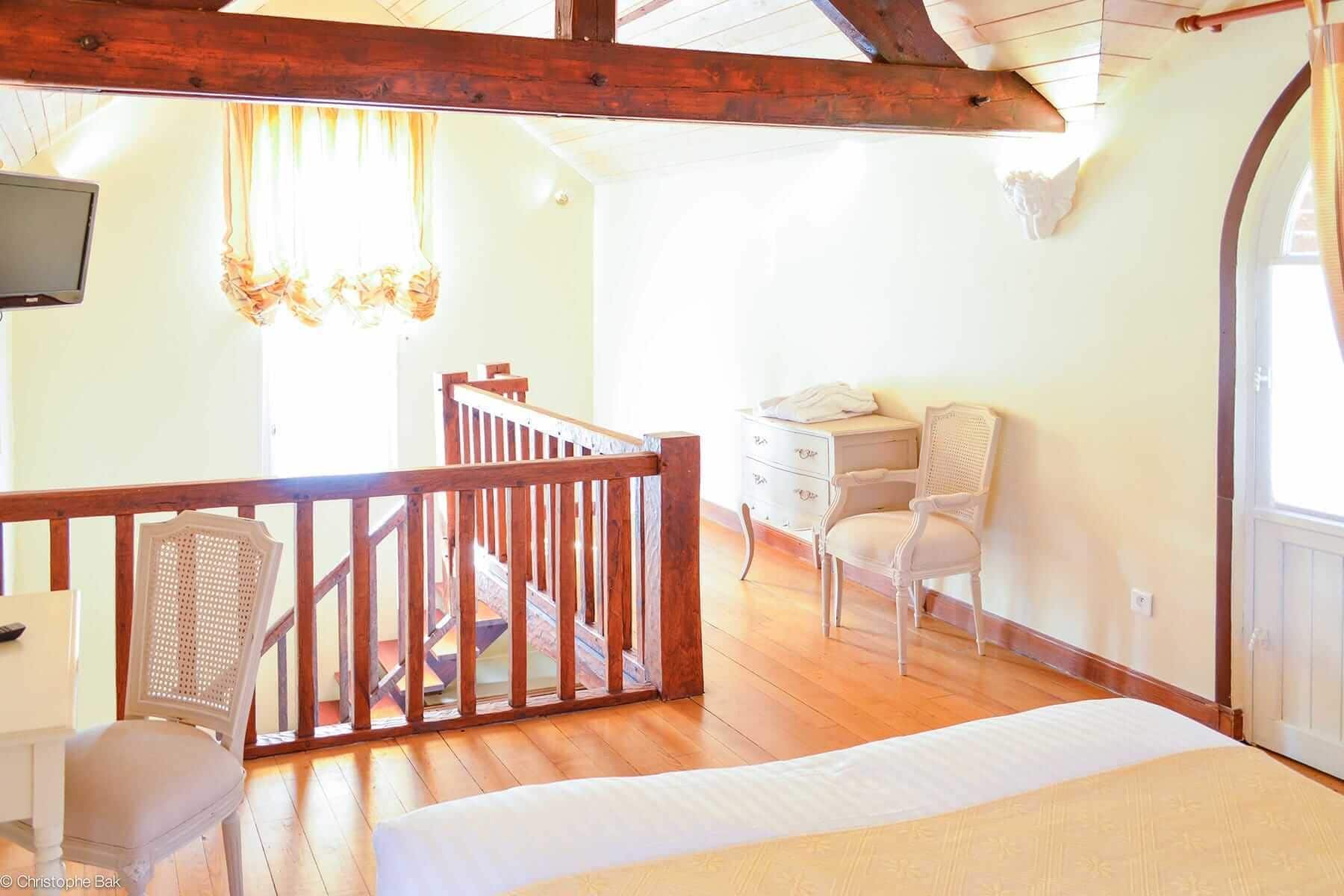 A bedroom with a wooden ceiling, a bed, a white dresser, a chair, and a staircase leading to an upper level.