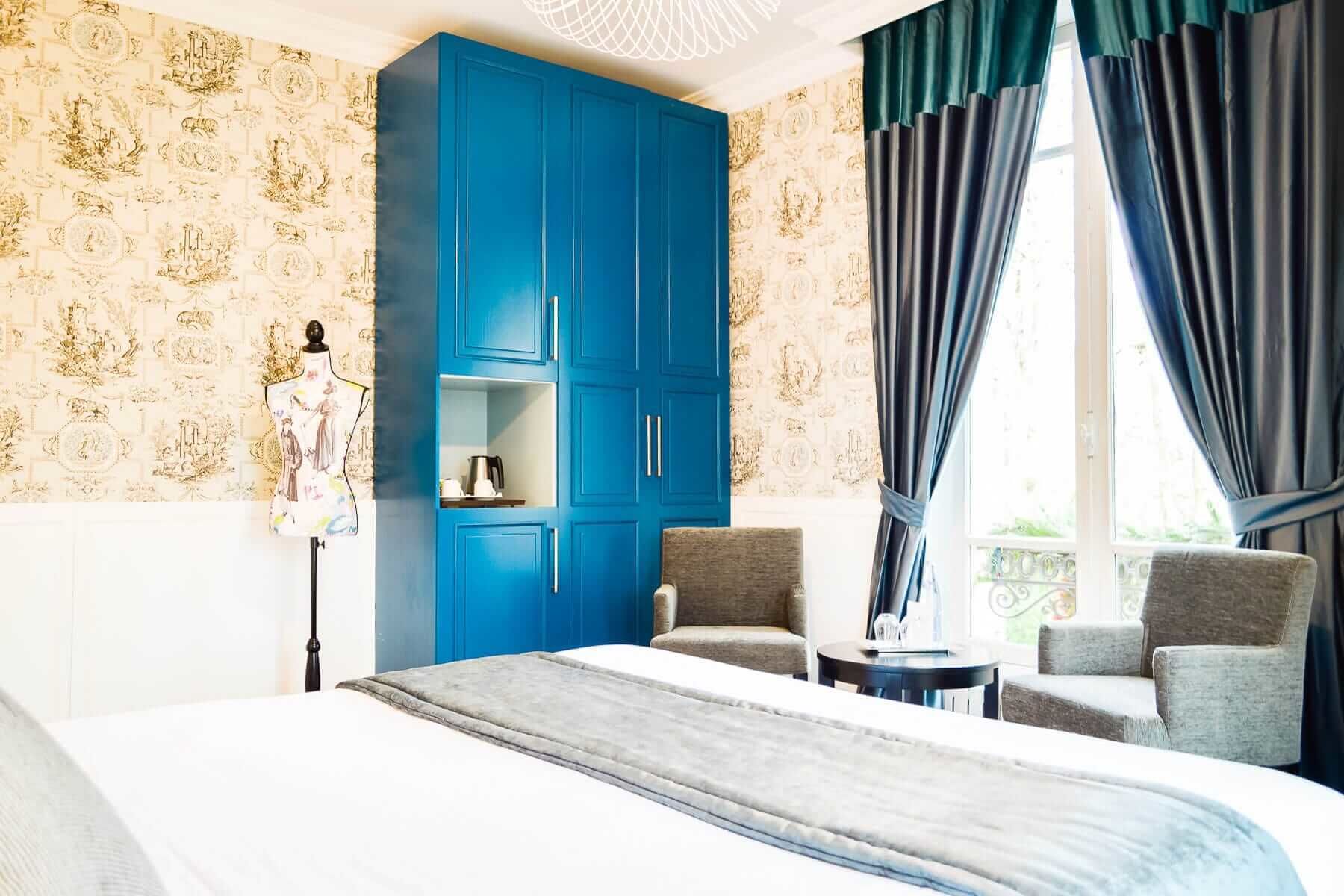 Une chambre d’hôtel avec un grand lit, une armoire bleue, deux fauteuils, une petite table et une fenêtre avec des rideaux.