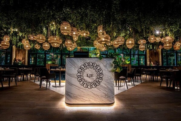 Interior de restaurante con mostrador de mármol, lámparas colgantes de mimbre y abundante vegetación.