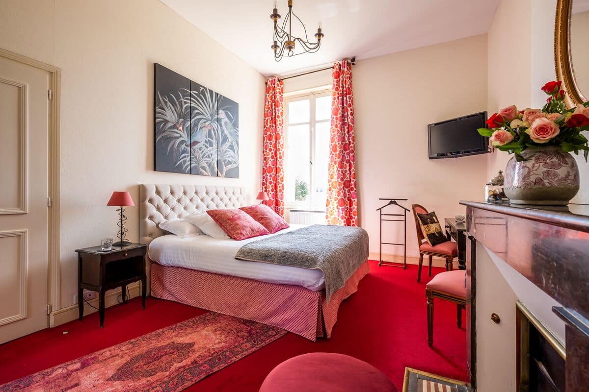 A bedroom with a tufted headboard bed, a red carpet, a large window with red curtains, a television, and a vase with flowers.