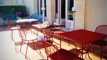 An outdoor patio with red metal tables and chairs, a blue chair, and a table set with a teapot, cups, and a plate with food.