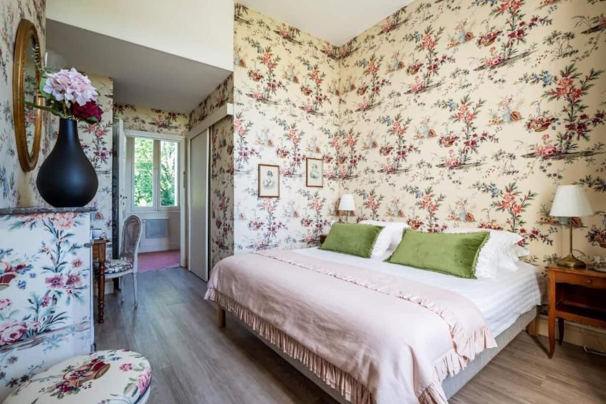 A bedroom with floral wallpaper, a bed with green pillows, a nightstand with a lamp, and a chair with floral upholstery.