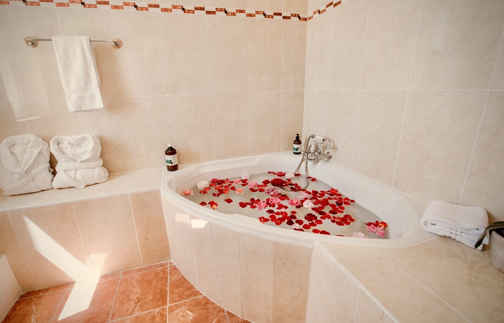 A bathroom with a bathtub filled with rose petals, a towel rack with towels, and a bottle of bath product.